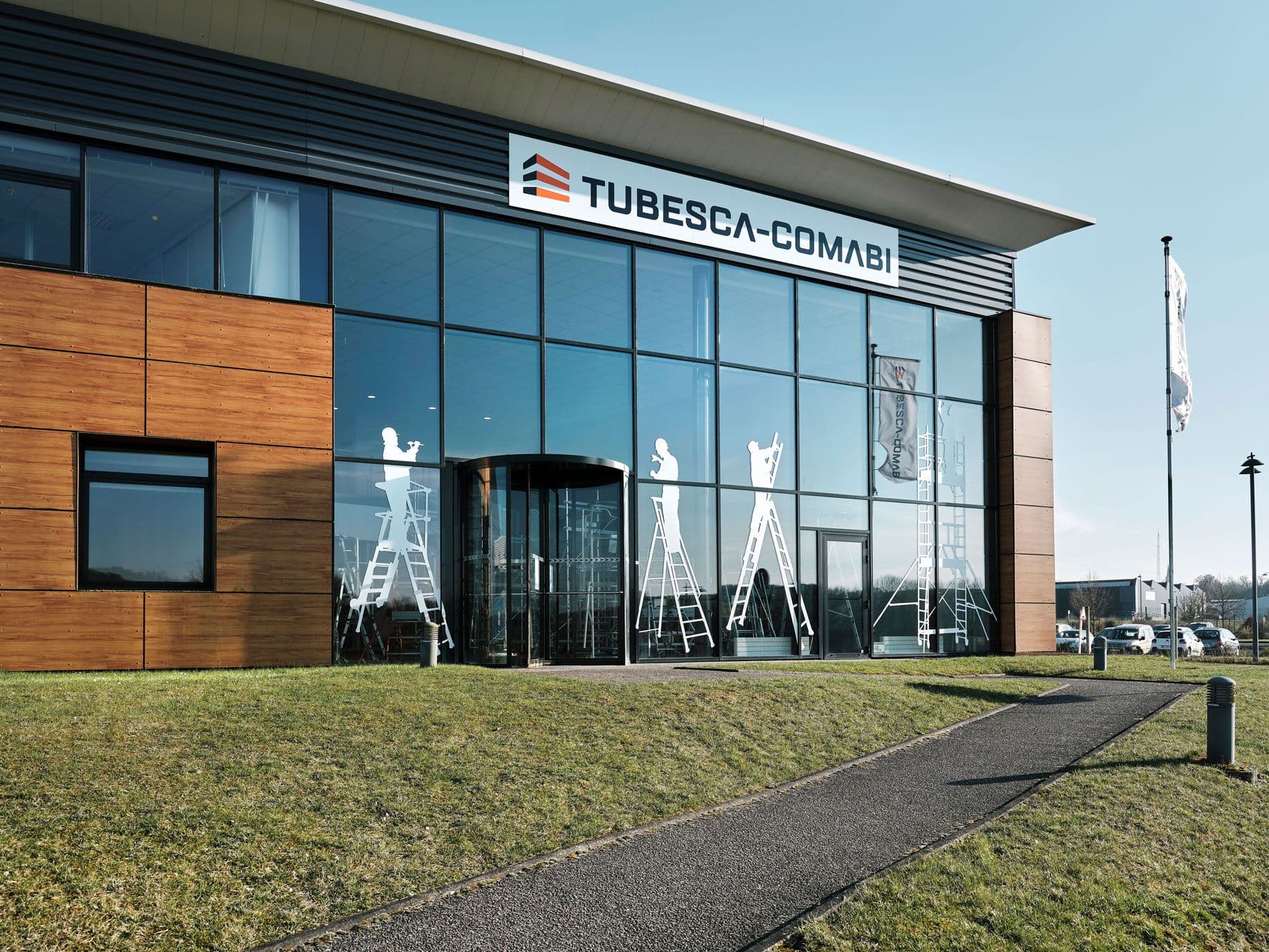 Modern two-story building with 'TUBESCA-COMABI' logo, glass facade, and wooden panels.